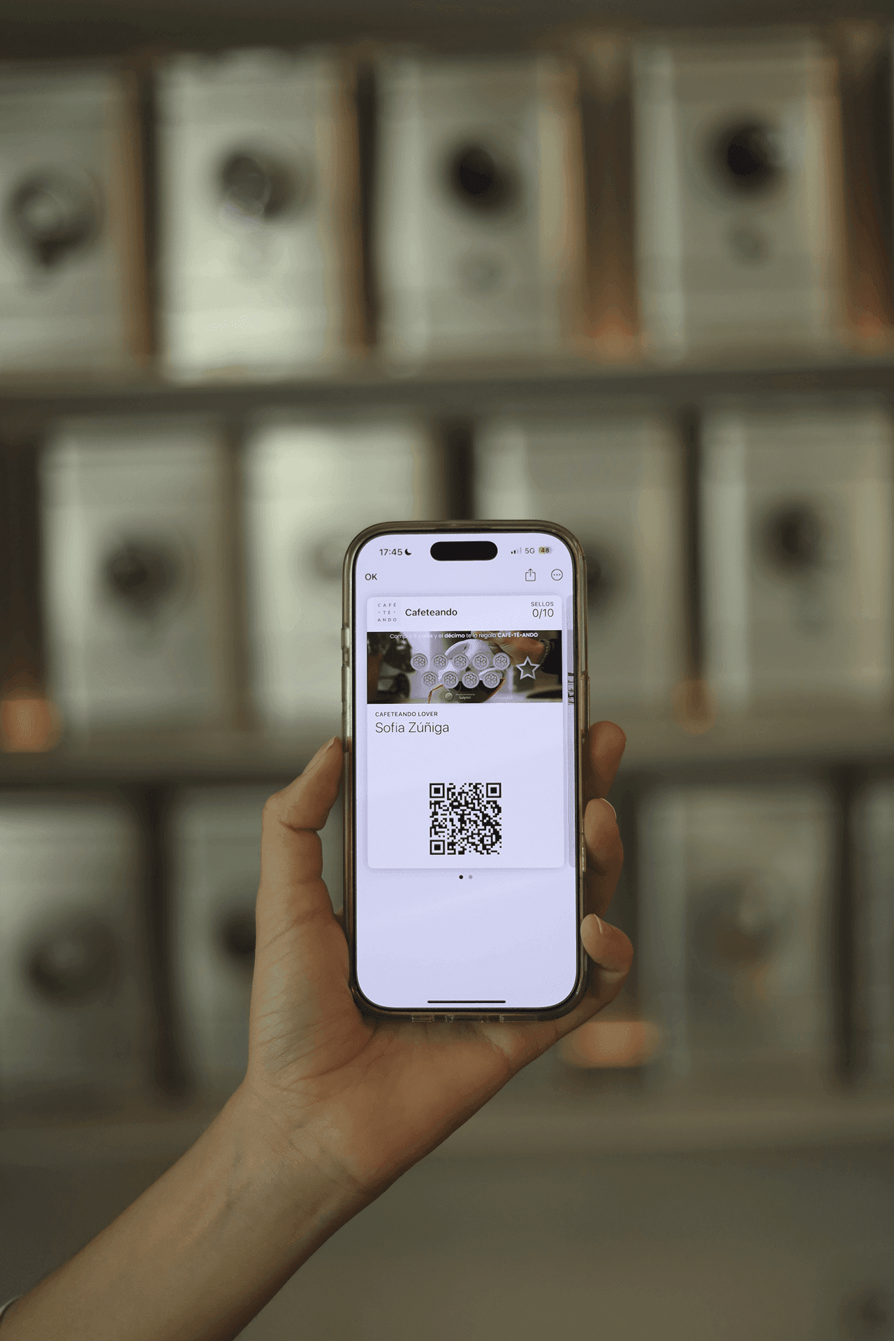 Mano sosteniendo un teléfono que muestra una tarjeta de lealtad digital con código QR creada por Luyoa en una cafetería